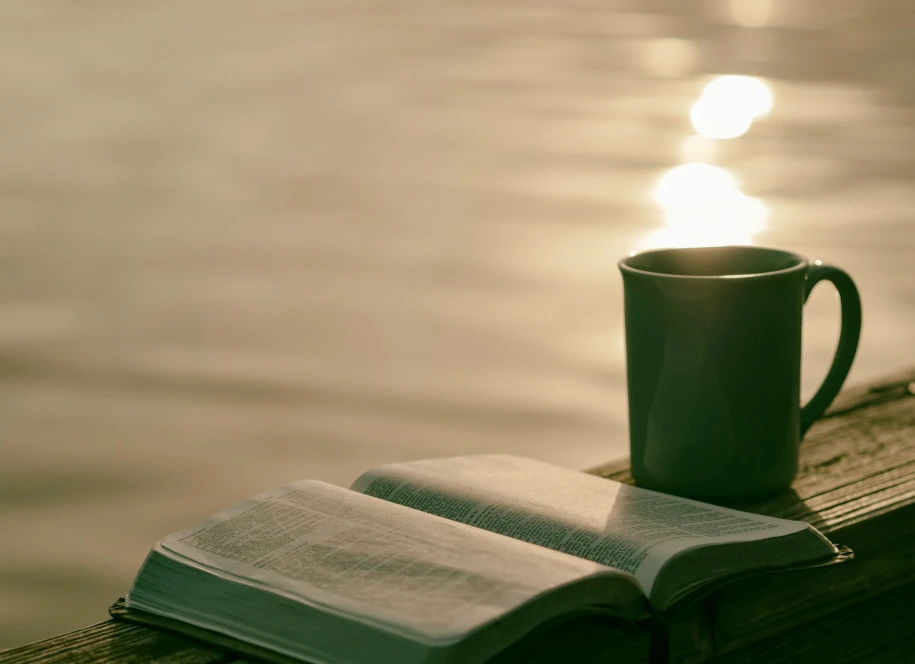 Une bible ouverte devant un plan d'eau au soleil couchnat avec une tasse en céramique verte à côté